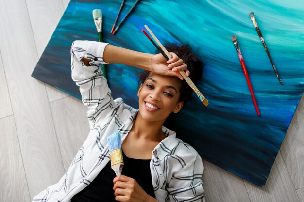 Top view of happy painter Africana American woman lying on canvas and looking at the camera with brushes in hands.