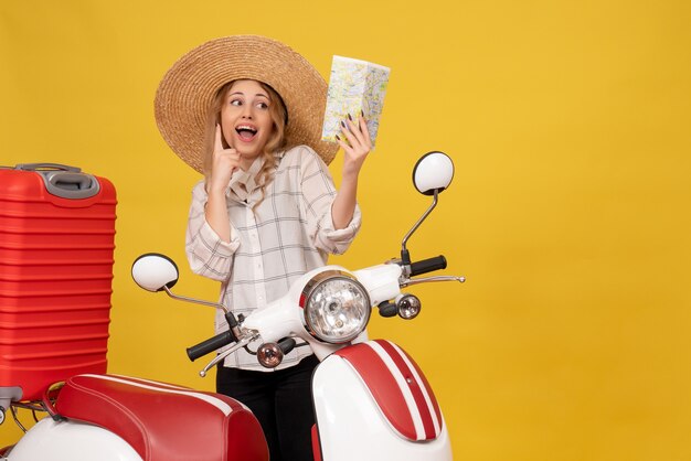 Top view of happy smiling young woman wearing hat collecting her luggage sitting on motorcycle and holding map