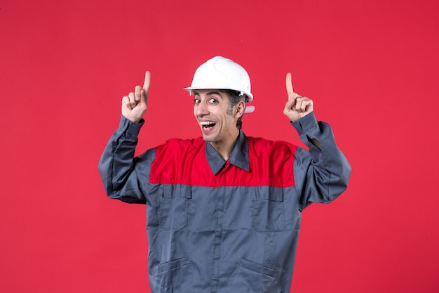 Top view of happy young builder in uniform wearing hard hat pointing up on isolated red wall