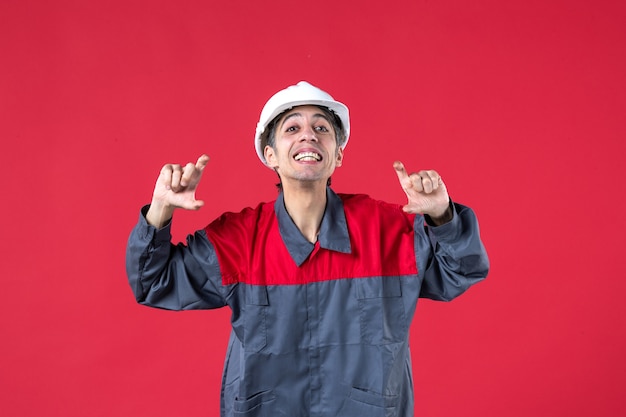 Foto gratuita vista dall'alto di un giovane costruttore felice in uniforme con elmetto che spiega qualcosa sul muro rosso isolato