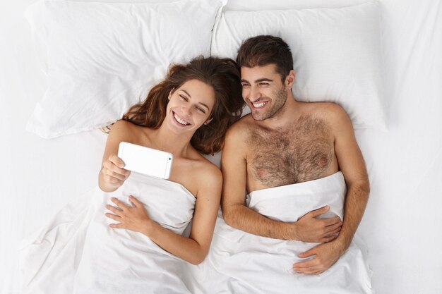 白いリネンのベッドで横になっていると携帯電話でselfieを取って幸せな若いヨーロッパのカップルの平面図