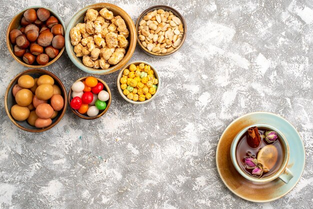 Top view of hazelnuts and peanuts with candies and cup of tea on white surface