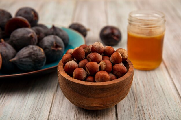 Top view of hazelnuts on a wooden bowl with ripe black mission figs on a blue dish with honey in a glass jar on a grey wooden wall