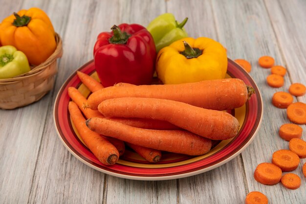 Top view of healthy bell peppers on a plate with carrots with yellow bell peppers on a bucket with chopped carrots isolated on a grey wooden background