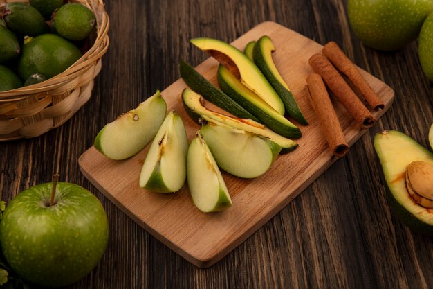 Вид сверху на здоровые нарезанные ломтики авокадо на деревянной кухонной доске с палочками корицы и ломтики яблока с фейхоа на деревянной поверхности