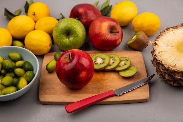 白い背景で隔離のカラフルなリンゴレモンとパイナップルとボウルにキンカンとナイフで木製のキッチンボード上のリンゴキウイスライスとキンカンなどの健康的な果物の上面図