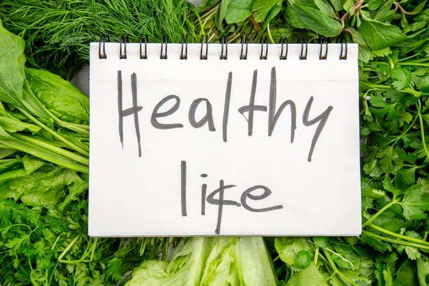 Top view of healthy life inscription on spiral notebook on bundles of fresh greens on white table