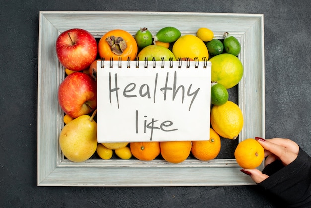 Top view of healthy life inscription on spiral notebook and hand taking one of tangerines from collection of fresh fruits in picture frame on dark background