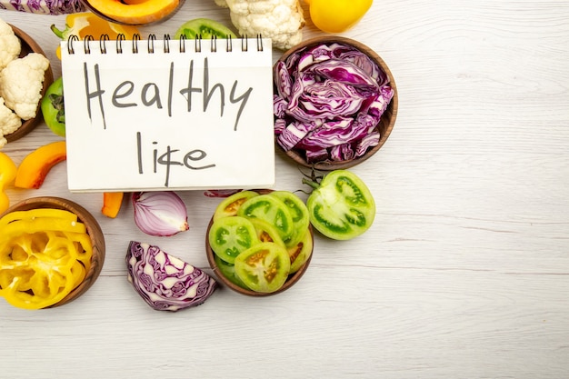Top view healthy life written on notebook cut green tomatoes cut red cabbage cut pumpkin cauliflower cut bell peppers in bowls on white wooden surface free space