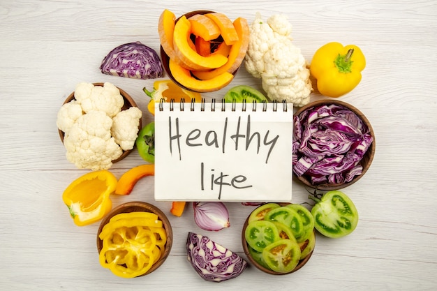 Top view healthy life written on notebook cut green tomatoes cut red cabbage cut pumpkin cauliflower cut bell peppers in bowls on wooden surface