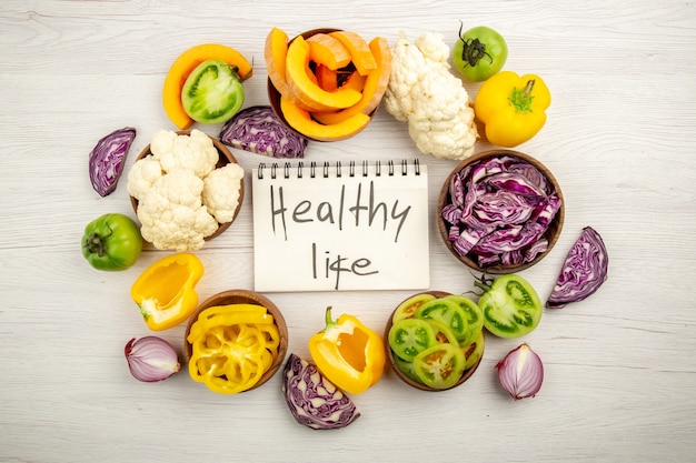 Top view healthy life written on notebook red cabbage cauliflower yellow bell pepper green tomato in bowls on surface