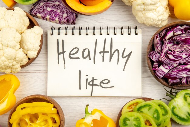Top view healthy life written on notebook red cabbage cauliflower yellow bell pepper green tomato in bowls on white wooden surface