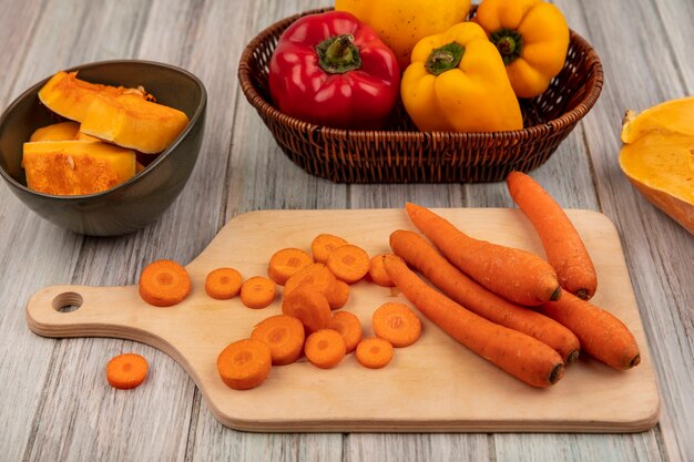 Top view of healthy orange carrots on a wooden kitchen board with colorful bell peppers on a bucket with pumpkin slices on a bowl on a grey wooden surface