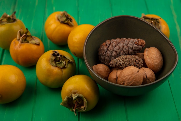 Вид сверху здоровых грецких орехов на миске с плодами хурмы, изолированными на зеленом деревянном столе