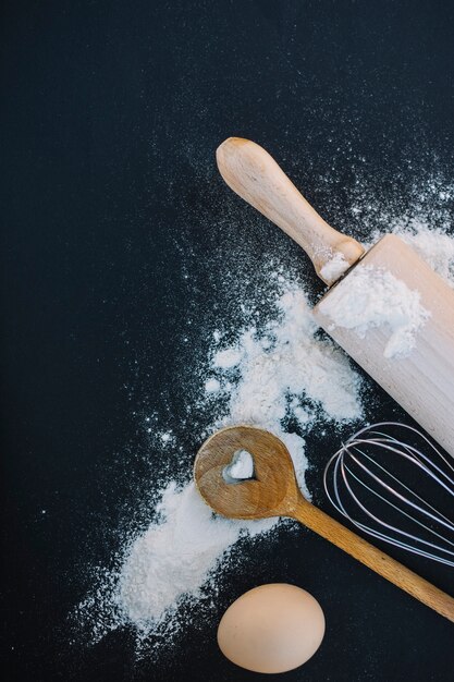 Top view of heart shape spoon; whisk; flour; egg and rolling pin on black background