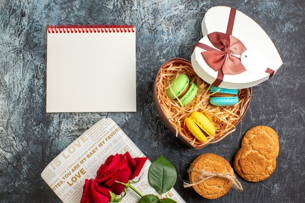 Top view of heart shaped beautiful gift box with delicious macarons and cookies red rose spiral notebook on icy dark background