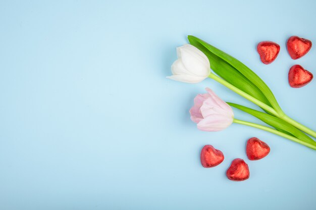 Top view of heart shaped chocolate candies in red foil with pink color tulips on blue table with copy space