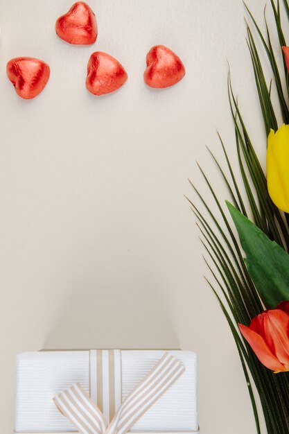 Top view of heart shaped chocolate candies wrapped in red foil, gift box and a bouquet of colorful tulips on white table with copy space