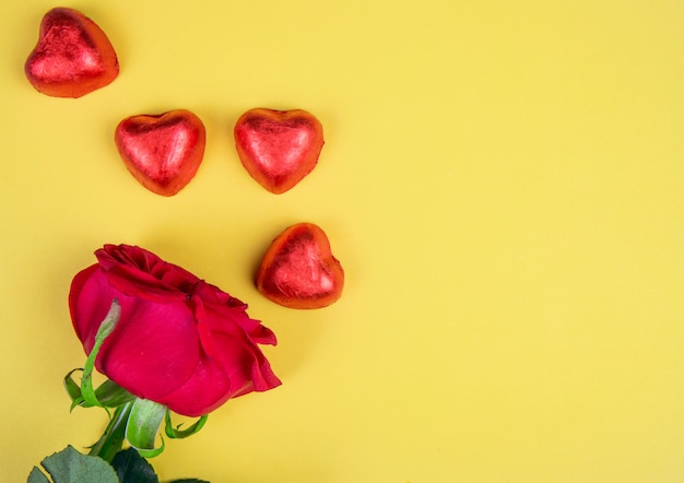 Top view of heart shaped chocolate candies wrapped in red foil with red rose on yellow table with copy space