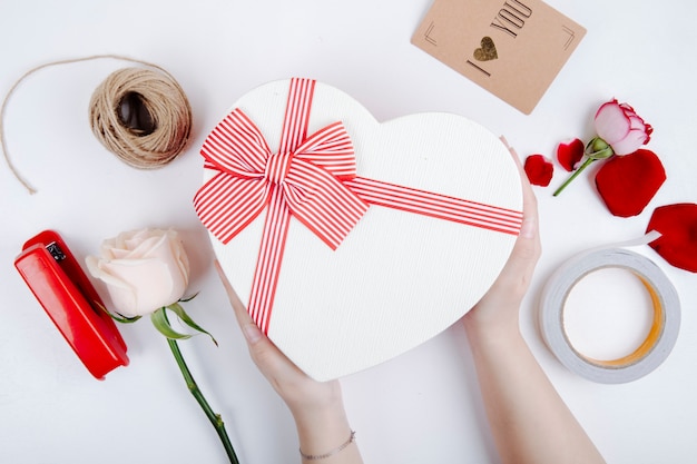 Top view of a heart shaped gift box with a bow and white color rose and stapler with rope and postcard on white background