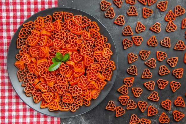 Top view heart shaped red italian pasta on black oval plate on kitchen towel scattered red heart pasta on dark table