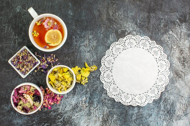 Top view of herbal tea near bowls of dry flowers with a piece of white lace on grey ground