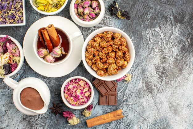 Top view of herbal tea with a bowl of nut and chocolate and different types of flower on grey ground