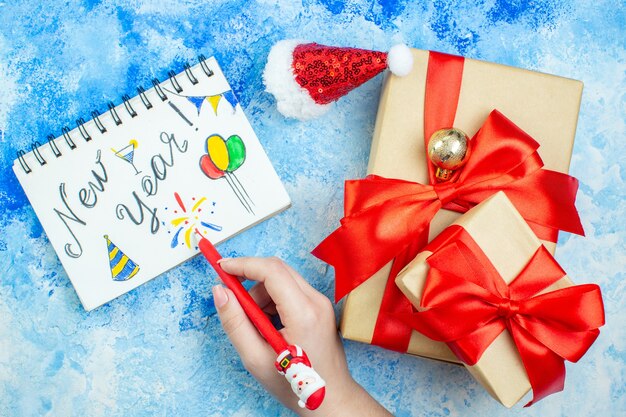 Top view holiday gifts small santa hat new year written on notepad pen in female hand on blue white table