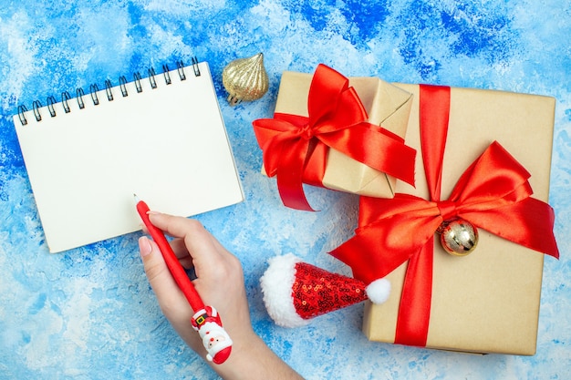 Foto gratuita regali di festa vista dall'alto piccolo blocco note cappello di babbo natale penna rossa di babbo natale in mano femminile sul tavolo bianco blu