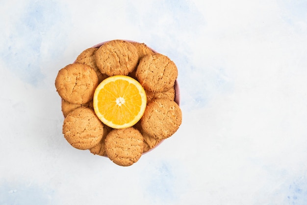 Top view of homemade cookies with half cut orange in bowl over white table.