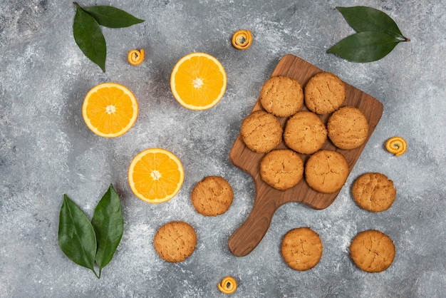 Top view of homemade cookies on wooden chopping board and half cut oranges with leaves.