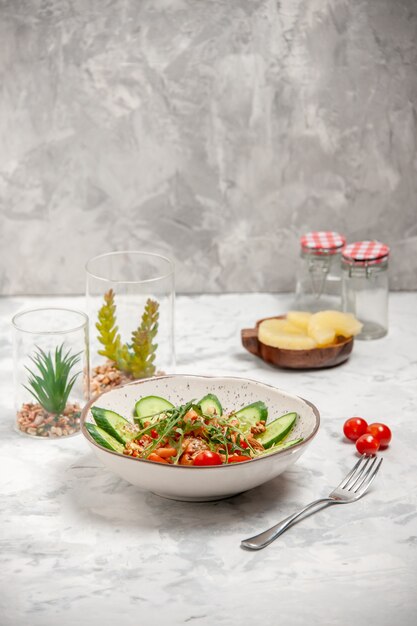 Вид сверху на домашний здоровый вкусный веганский салат, украшенный нарезанными огурцами в миске, вилкой, сушеными ананасами, помидорами на окрашенной белой поверхности