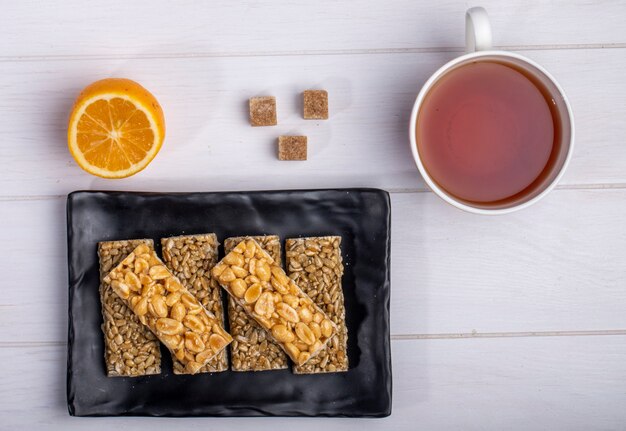 Top view of honey bars with peanuts and sunflower seeds on a black platter with a cup of tea and lemon on white