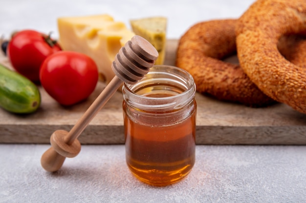 Вид сверху на мед в стеклянной банке с деревянной ложкой для меда с турецкими бубликами, сырными огурцами и помидорами на деревянной кухонной доске на белом фоне