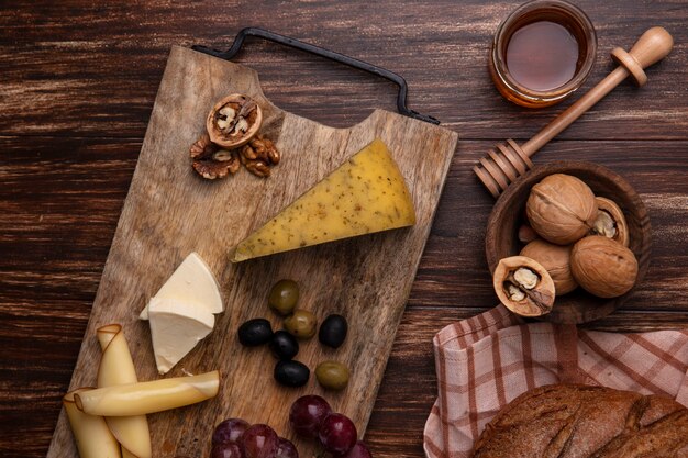 Top view honey in a jar with walnuts  and a loaf of black bread with varieties of cheeses and grapes on a stand  on a wooden background