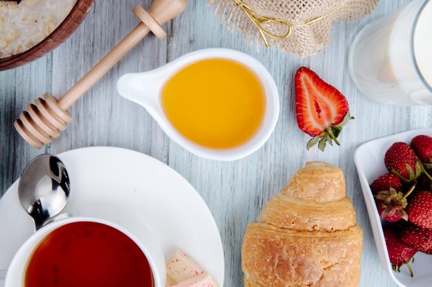 La vista superiore di miele in un piattino con le fragole mature fresche del croissant è servito con una tazza di tè su rustico