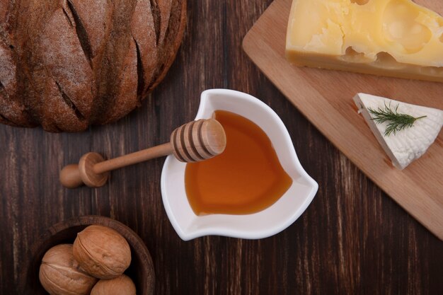 Top view honey in a saucer with a variety of cheeses on a stand with walnuts and a loaf of bread on a wooden background