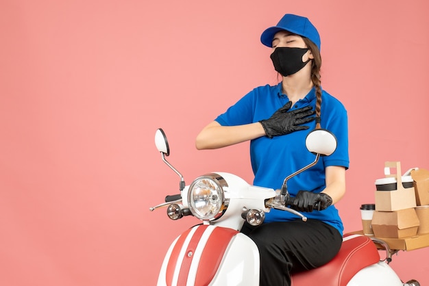 Top view of hopeful courier girl wearing medical mask and gloves sitting on scooter delivering orders on pastel peach background