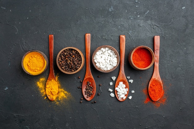 Top view horizontal row bowls with turmeric black pepper sae salt red pepper powder wooden spoons on black table