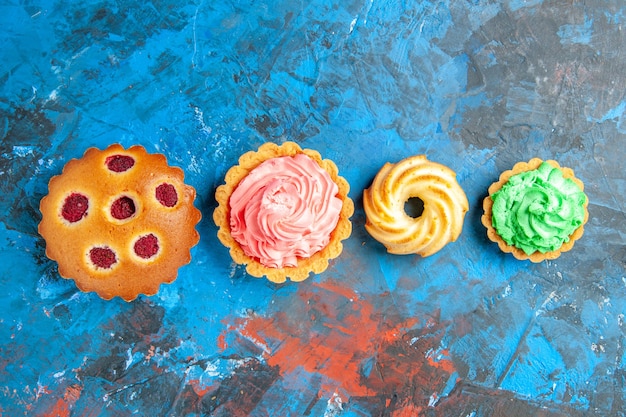 Top view of horizontal row raspberry cupcakes, small tarts and biscuit on blue surface