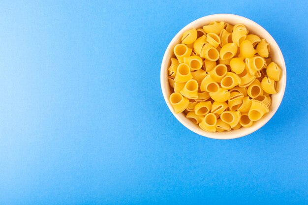 トップビューイタリアの乾燥パスタは青い背景のイタリアのスパゲッティ食品パスタに分離されたクリーム色の丸いボウルの中に少し黄色の生パスタを形成