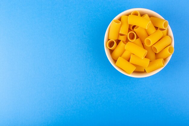 トップビューイタリアの乾燥パスタは、青に分離されたクリーム色の丸いボウルの中の小さな黄色の生パスタを形成
