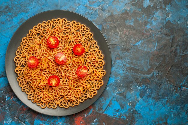 상위 뷰 이탈리아 파스타 하트는 복사 장소가 있는 파란색 테이블에 있는 검은 타원형 접시에 체리 토마토를 잘라냅니다.