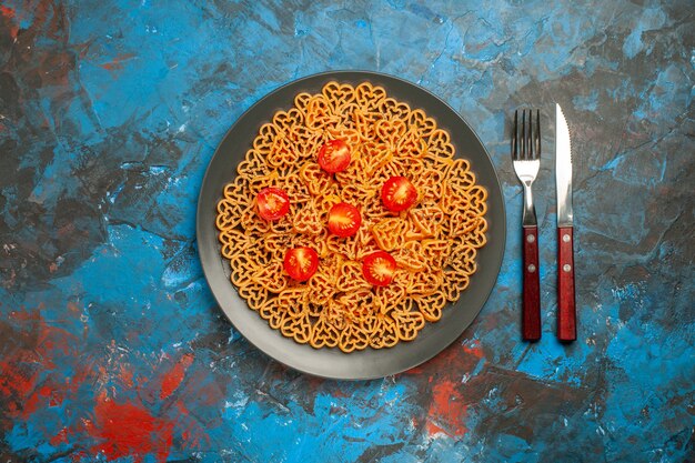 Top view italian pasta hearts cut cherry tomatoes on black oval plate fork and knife on blue table copy place