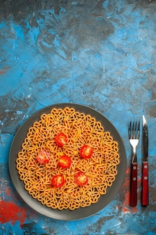 Vista dall'alto cuori di pasta italiana tagliati pomodorini su forchetta e coltello piatto ovale nero sul tavolo blu con spazio libero
