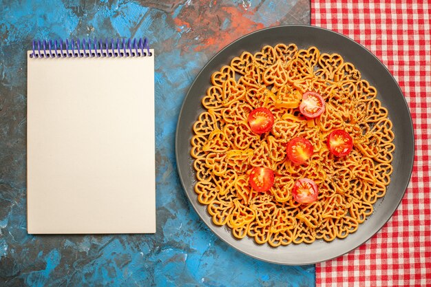 Foto gratuita vista dall'alto cuori di pasta italiana tagliati pomodorini su piatto ovale su quaderno tovaglia a quadretti bianchi rossi sul tavolo blu