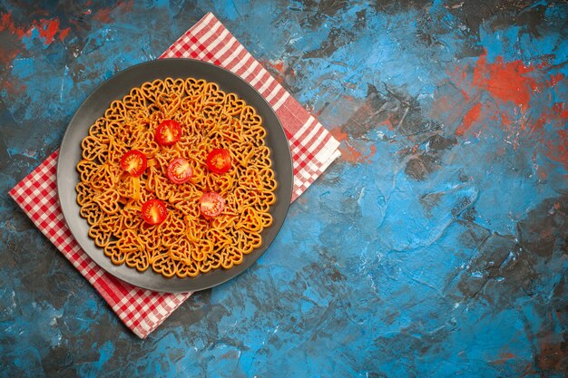 상위 뷰 이탈리아 파스타 하트는 여유 공간이 있는 파란색 테이블에 있는 빨간색 흰색 체크 무늬 주방 수건에 접시에 체리 토마토를 잘라냅니다.