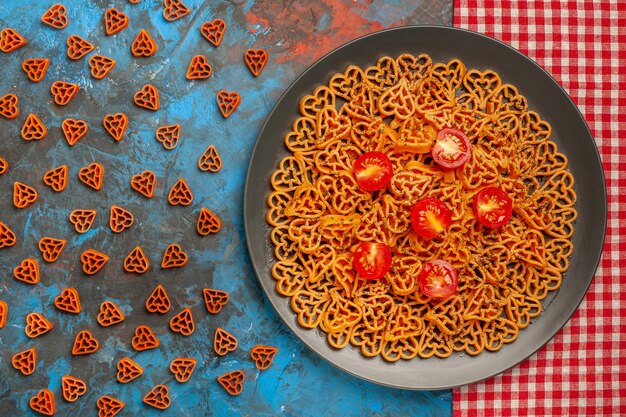 Top view italian pasta hearts cut cherry tomatoes on plate on red white checkered tablecloth scattered red heart pasta on blue table