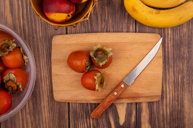 Vista dall'alto di succosi cachi su una tavola da cucina in legno con coltello con pesche su un secchio con banane isolato su uno sfondo di legno