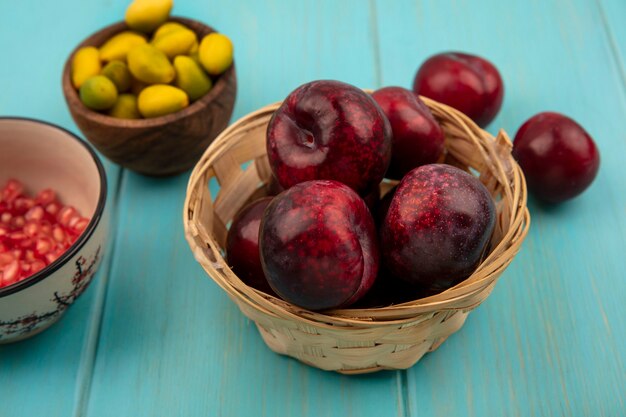 Top view of juicy pluots on a bucket with pomegranate seeds on a bowl with kinkans on a wooden bowl on a blue wooden background